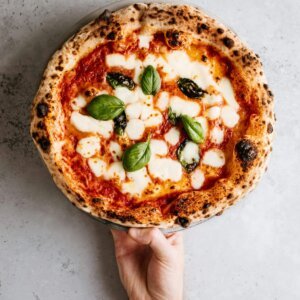 Close-up of a fresh Margherita pizza with mozzarella and basil, held over a gray background.