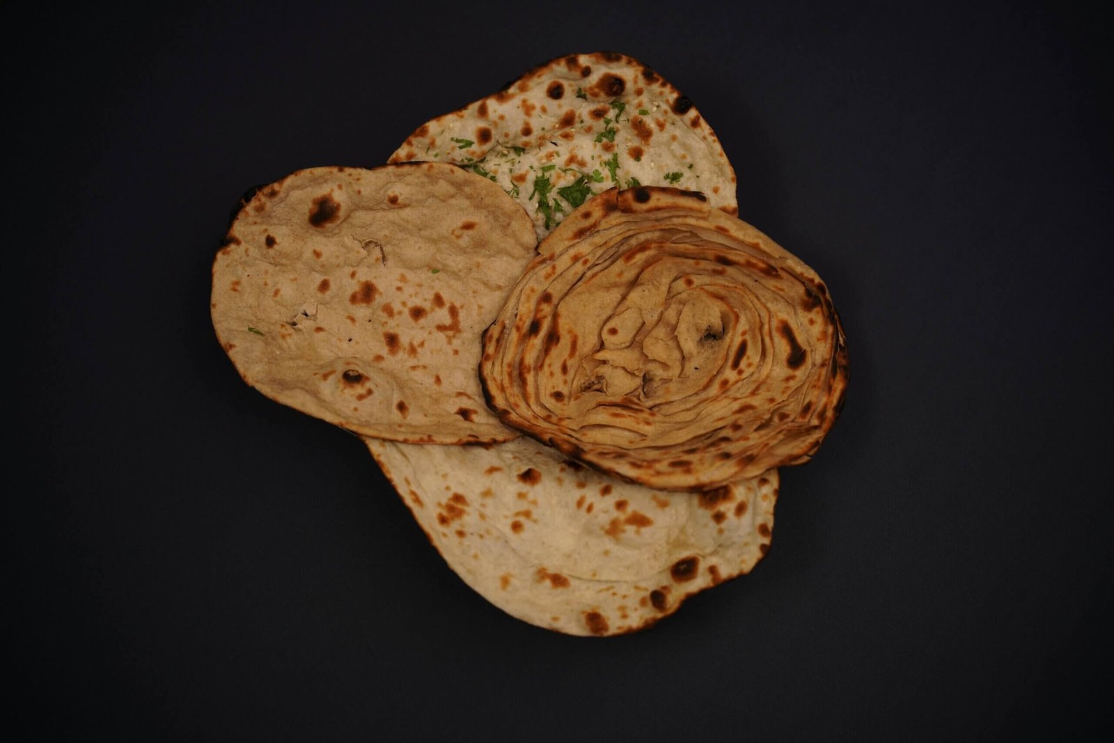 Top-down view of various Indian breads including lachha paratha and naan on a dark background.